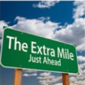 Square badge with a green road sign reading The Extra Mile Just Ahead
